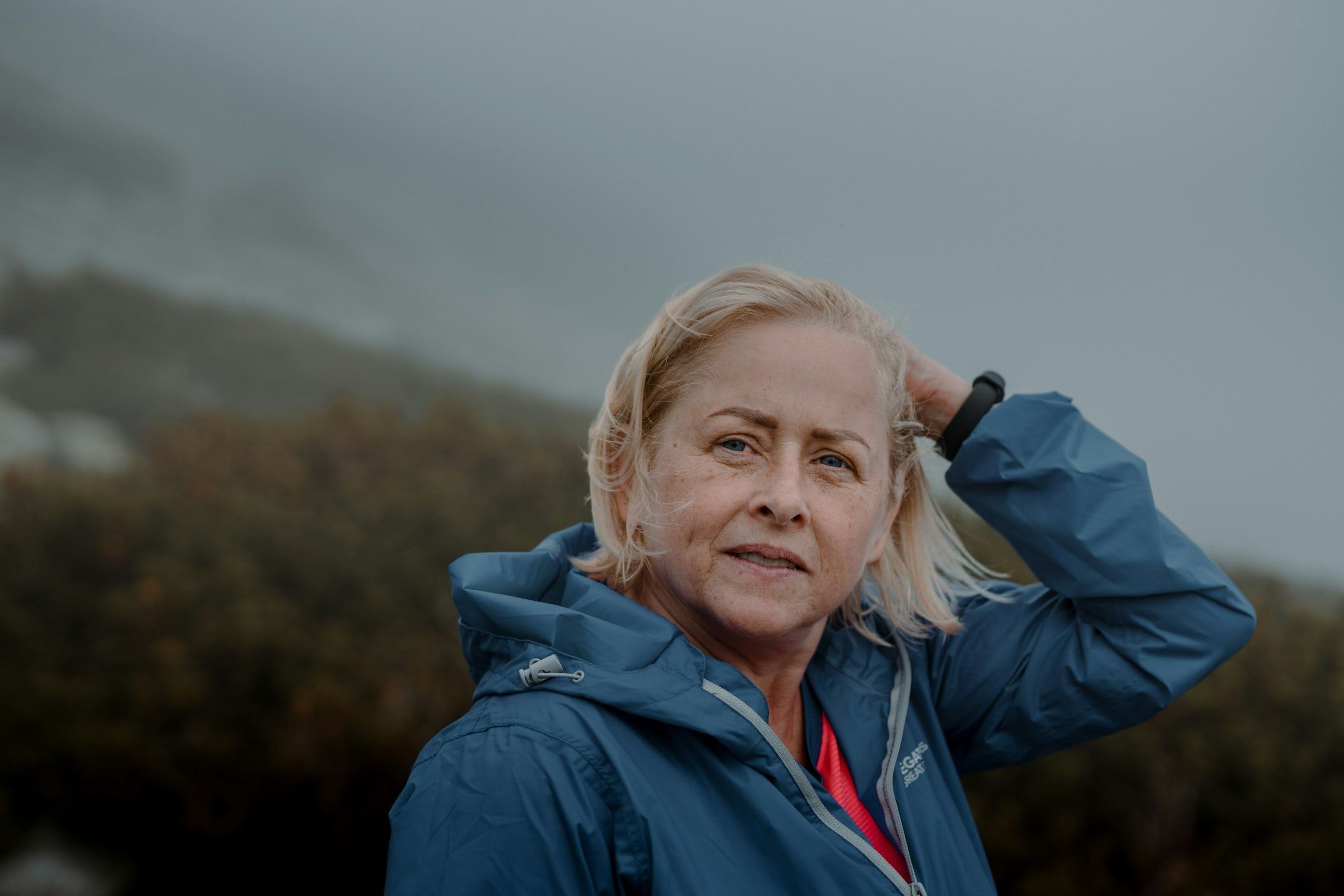 An older woman outdoors in a blue jacket, wind in her hair, looking toward the camera.
