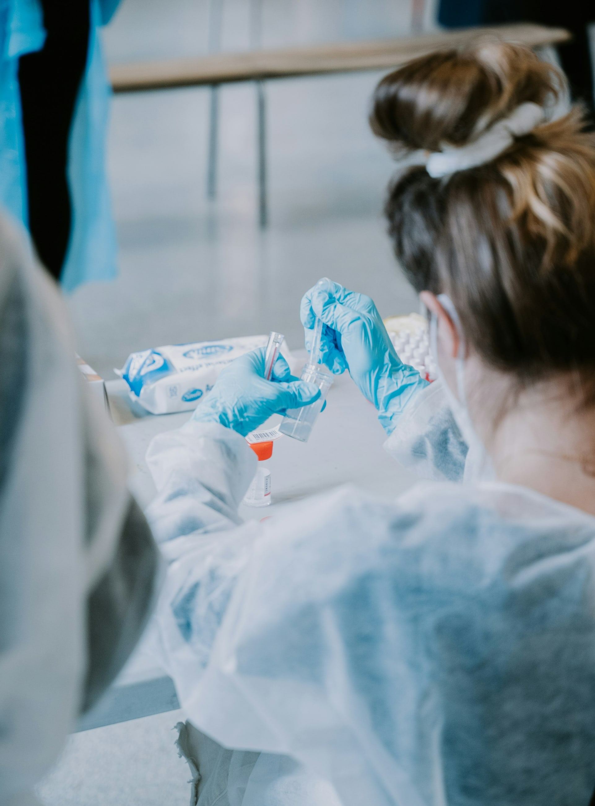 A health worker in a protective gown, mask and gloves preparing a sample tube at a testing station.